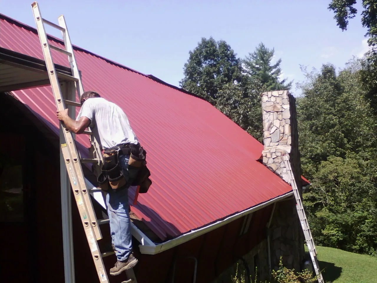 Roofer on a ladder beside a red metal roof and stone chimney — Soffit & Fascia Repair by Roofing Brothers in Winchendon