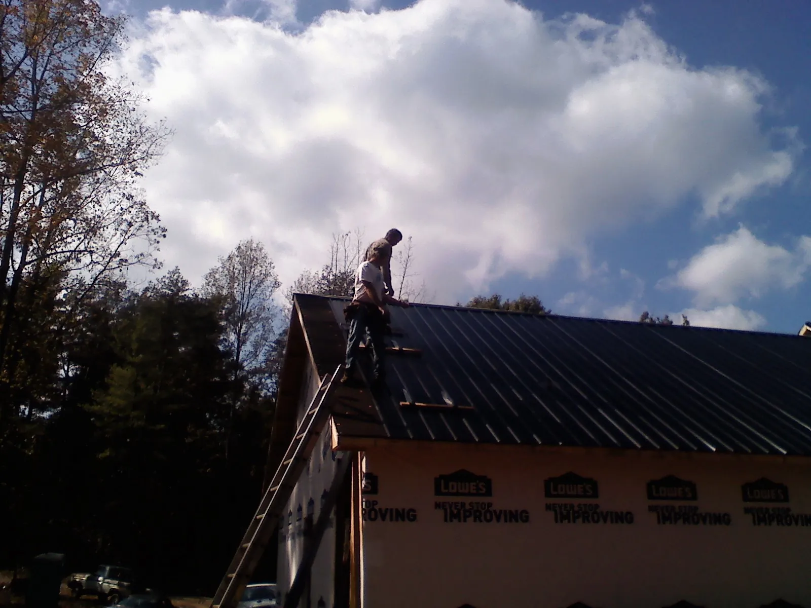 Roofer fastening dark metal panels high against a cloudy sky, from a Roof Inspection job in Winchendon